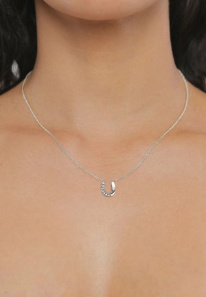 Zarte silberne Halskette mit einem kleinen Hufeisenanhänger, der mit fünf kleinen Kristallen verziert ist, getragen am Hals einer Frau.