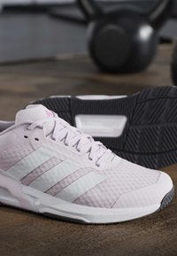 Ανοιχτό ροζ αθλητικό παπούτσι Adidas με λευκές ρίγες σε ξύλινο πάτωμα και θολό μαύρο kettlebell στο φόντο.