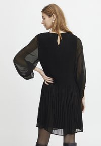 Schwarzes, plissiertes Kleid mit transparenten, langen Ärmeln, Rückendetail in Herzform und fließender Silhouette, das einen strukturierten Oberteil und einen glatten Stoff aufweist.
