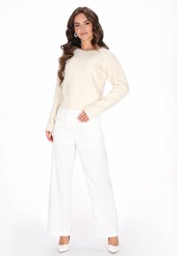 Suéter de punto crema con mangas largas combinado con pantalones blancos de talle alto y pierna ancha. Modelo llevando tacones altos blancos y pendientes.