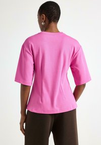 Rosa överdimensionerad t-shirt med korta ärmar, med rund halsringning och detaljer i sömmen på baksidan, tillsammans med bruna byxor. Slät bomullstextur.