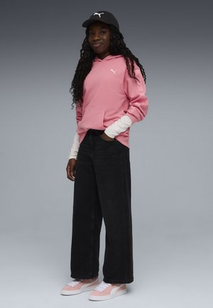 Jeune personne portant un sweat à capuche rose Puma, un tee-shirt à manches longues blanc en dessous, un pantalon ample noir, des baskets roses et blanches, et une casquette noire Puma.