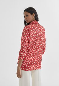 Camicia rossa con un motivo geometrico bianco, chiusura con bottoni sul retro, colletto e maniche arrotolate, realizzata in tessuto leggero. Indossata con pantaloni bianchi.