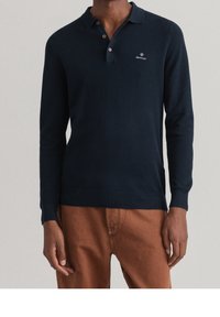 Navyblaues Langarm-Polohemd mit strukturiertem Stoff, zwei Knöpfen am Ausschnitt und gesticktem Logo auf der linken Brust, kombiniert mit beigen Hosen.