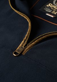 Marineblauwe fleece hoodie met een bruin rits met gouden hardware. Gedetailleerde stiksels en een label met de merknaam 'Superdry' zichtbaar.