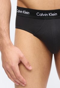 Férfi törzs és kéz fekete Calvin Klein alsóneműben, márkás derékrésszel, világos háttéren.