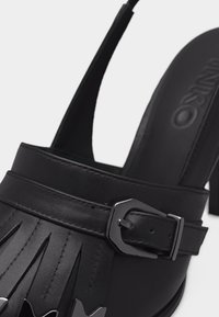 Chaussure slingback en cuir noir avec une boucle argentée et des détails à franges sur la partie supérieure.