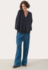 Marineblauwe oversized breipullover met een V-hals en verlaagde schouders, gecombineerd met lichtblauwe denim met wijde pijpen en bruine schoenen.