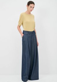 Top beige a maniche corte abbinato a pantaloni in denim scuri a gamba larga, con vita alta, pieghe e tasche laterali. Linee pulite e dettagli minimi.