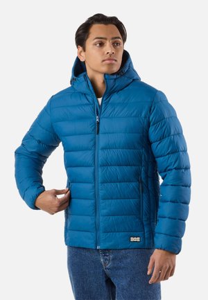 Young man wearing a blue quilted hooded jacket with front zipper and side pockets, adjusting zipper on the left pocket, against white background.