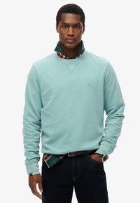 Licht turkoois sweatvest met ribbelboorden bij de mouwen en de hals, gelaagd over een geblokt shirt in kleuren groen, rood en marineblauw. Donkere jeans en een horloge.