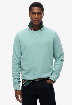Licht turkoois sweatvest met ribbelboorden bij de mouwen en de hals, gelaagd over een geblokt shirt in kleuren groen, rood en marineblauw. Donkere jeans en een horloge.