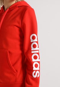 Felpa rossa con cappuccio realizzata in stoffa morbida, dotata di una grande tasca frontale e del logo "adidas" stampato in bianco in verticale sulla manica sinistra.