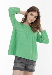 Groene gebreide trui met een getextureerd chevronpatroon, ronde halslijn en lange mouwen, gecombineerd met grijze shorts.
