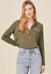 Camisa de manga larga verde olivo con cuello, que presenta dos bolsillos en el pecho y un corte relajado, combinada con jeans de mezclilla claros de tiro alto.