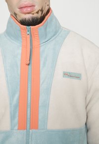 Giacca in fleece Columbia con chiusura a zip, caratterizzata da blocchi di colore azzurro chiaro, beige e corallo. Tessuto testurizzato e patch con logo presente.
