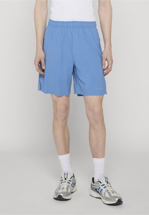 Mees, kes kannab helesiniseid sportlühikondseid, valgeid sokke ja valgeid jooksujalatseid siniste detailidega, seisab ühevärvilise tausta ees.