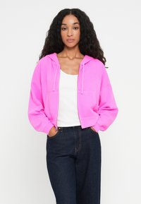 Hoodie fucsia cropped con cappuccio regolabile, dotato di tasche frontali e morbida texture, abbinato a jeans blu scuro.