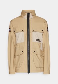 Giacca beige con colletto alto, due tasche sul petto e due tasche inferiori. Presenta una zip nera e una toppa con logo sulla manica.