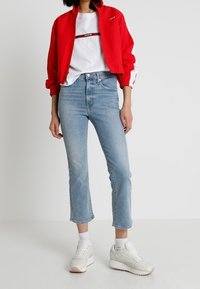 Czerwona bomberka z białymi akcentami, biała koszulka, niebieskie skrócone dżinsy i białe trampki z jasnoróżowymi podeszwami.