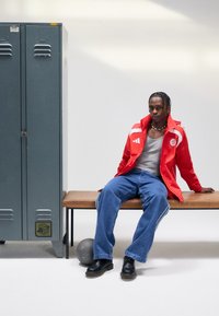 Mladá osoba v červenej športovej bunde a modrých džínsoch sedí na lavičke vedľa sivých skríň, pričom na podlahe sa nachádza basketbalová lopta.