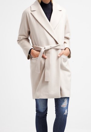 Mujer con un abrigo beige con cinturón sobre un jersey de cuello alto oscuro y vaqueros azules rotos, manos en los bolsillos del abrigo, sobre un fondo blanco.