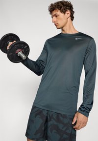 Langærmet, mørk teal sportstrøje med glat overflade, prydet med et lille hvidt Nike-logo på brystet. Holder en sort håndvægt.