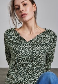 Blusa verde a fantasia con scollatura a V e collo arricciato, caratterizzata da un design geometrico nei colori verde scuro e crema. Tessuto testurizzato.