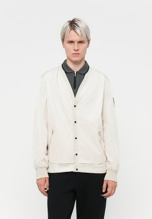 Light beige bomber jacket with a zip-up collar, snap buttons, and side pockets. Features elastic cuffs and a smooth texture.
