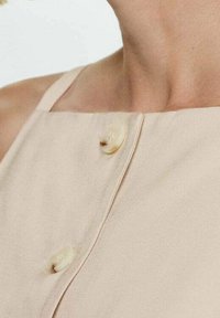 Haut beige sans manches avec une texture lisse, doté d'une fermeture à bouton et d'un design simple, mettant en valeur la qualité du tissu et ses lignes épurées.