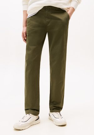 Άτομο που φοράει χακί παντελόνι με ίσια γραμμή, λευκά αθλητικά παπούτσια με μαύρες λεπτομέρειες και κρεμ πουλόβερ, όρθιο με τα χέρια στις τσέπες.