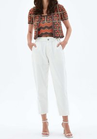 Mujer luciendo un top corto de crochet en tonos marrón y naranja con un cárdigan corto a juego, pantalones blancos de talle alto y sandalias de tacón alto blancas.