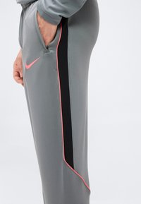 Άτομο που φοράει γκρι φόρμα της Nike με μαύρη ρίγα στο πλάι και ροζ περιθώριο, με το χέρι εν μέρει στην τσέπη με φερμουάρ.