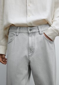Person iført en lysebeige knapbluse, der er stoppet i lysegrå jeans, med højre hånd i lommen og venstre hånd afslappet ved siden.