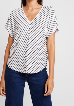 Femme portant un haut blanc à manches courtes et encolure en V avec des rayures diagonales noires, associée à un jean bleu foncé, debout devant un fond uni.