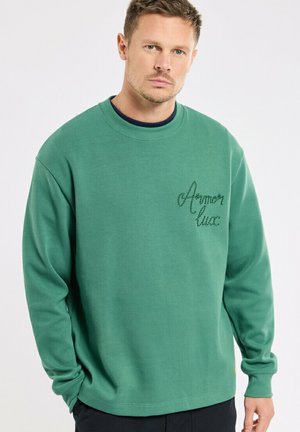 Homme portant un sweat-shirt vert à manches longues avec le texte brodé "Aymor luzc" sur la poitrine, debout devant un fond uni.