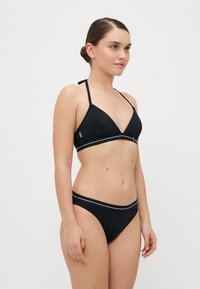 Melna bikini komplekts, kas satur trīsstūra formas augšdaļu ar plānām siksnām un zemu jostasvīru apakšdaļu, ko akcentē baltas horizontālas svītras visā joslā.