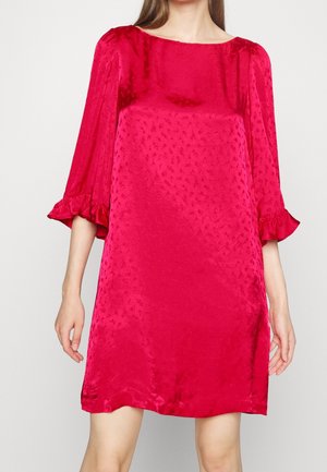 Femme portant une robe ample en satin rouge vif, avec des manches trois-quarts et des poignets à volants, debout devant un fond uni.