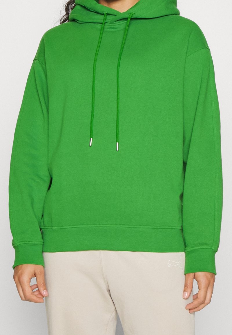 Personne portant un sweat à capuche vert vif avec des cordons, associé à un pantalon beige clair, debout devant un fond uni.