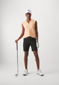 Polo tričko s krátkymi rukávmi v broskyňovej farbe s golierom, skombinované s čiernymi šortkami. Súčasťou outfitu je biela čiapka a golfová palica. Na ľavej ruke je nasadená textúrovaná golfová rukavica.