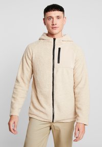 Beige fleece hoodie med lynlås foran, sort lynlås og en ensfarvet panel på brystet. Blød tekstur og lange ærmer.