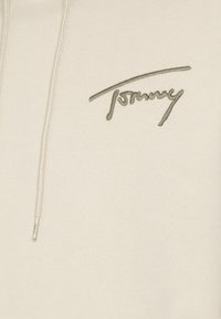 Světle béžová mikina s hladkou texturou, na přední straně s vyšitým zeleným logem "Tommy" a zipem s kovaným stahovákem ve stříbrné barvě.