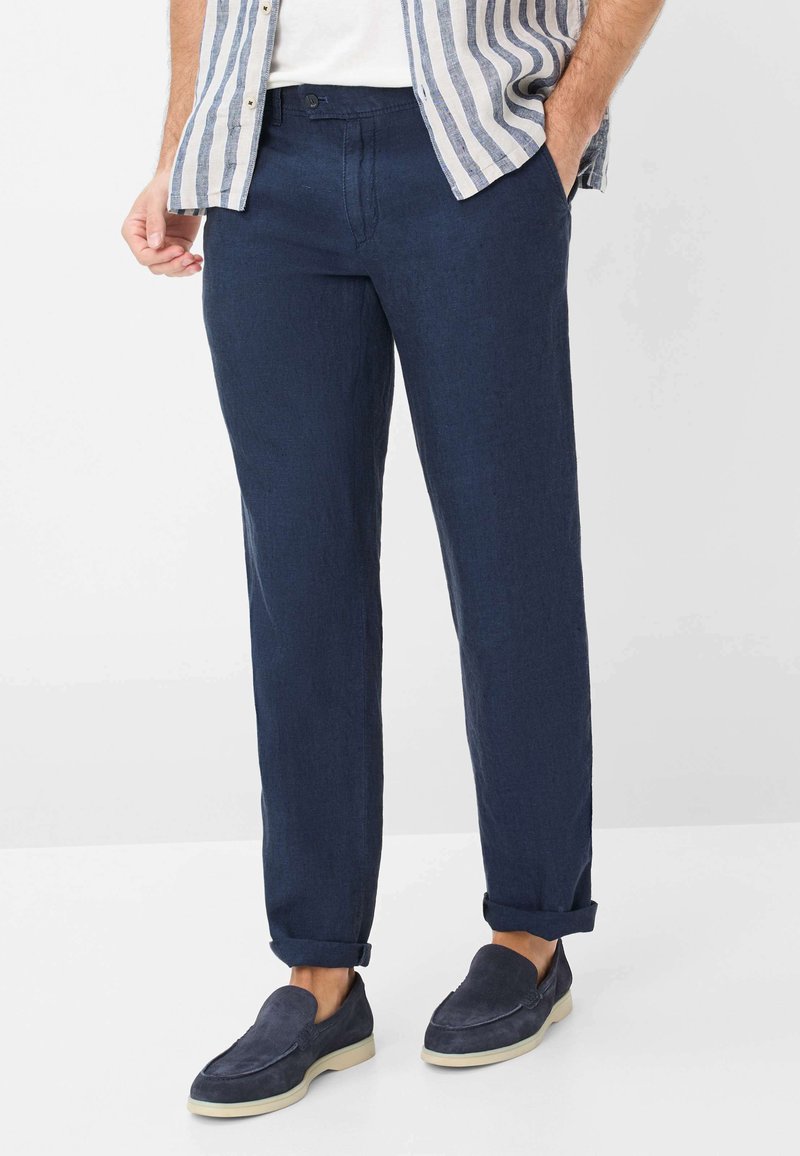 Pantalons en lin bleu marine pour homme avec une coupe décontractée. Caractéristiques d'une texture lisse, de ourlets retroussés et de plis avant subtils. Portés avec des chaussures slip-on bleu marine.