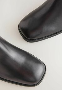Botas de cuero negras hasta el tobillo con un acabado suave. Diseño de punta cuadrada, costuras en contraste y una suela plana y texturizada.