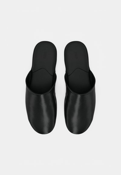 Une paire de pantoufles noires en cuir, à enfiler, avec des bouts fermés et des talons ouverts, au design minimaliste, présentée sur fond blanc.