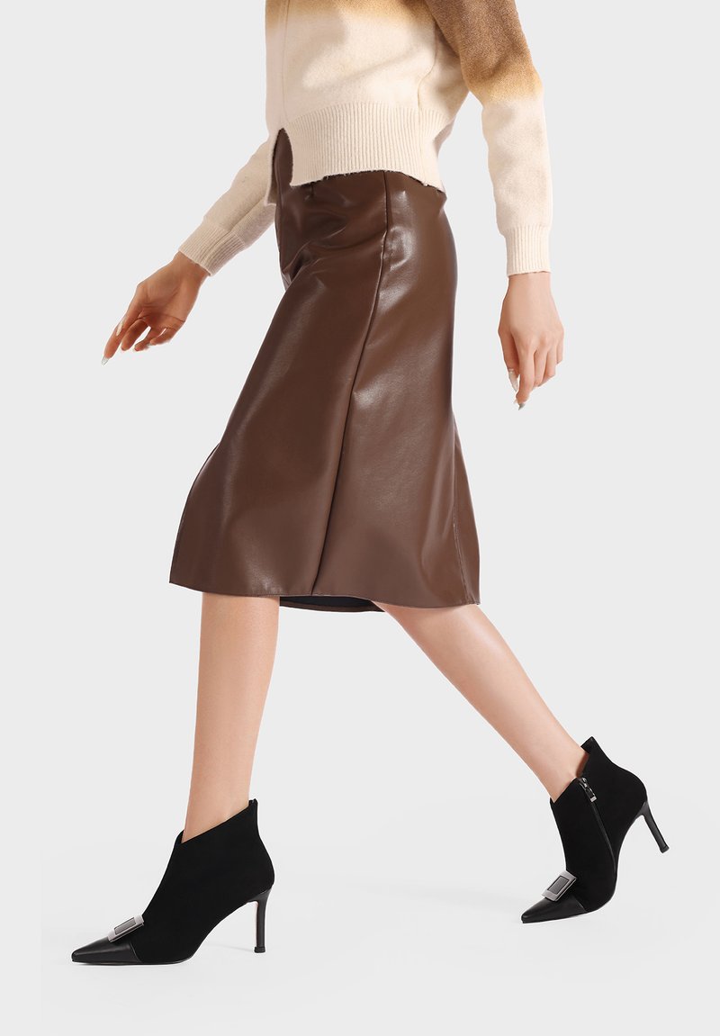 Gonna midi in finta pelle marrone abbinata a stivaletti neri in suede alla caviglia con punta affusolata e dettagli metallici. Modella in posa mentre cammina.