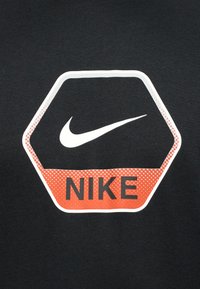 Εξαγωνικό λογότυπο της Nike με λευκή σημάδια πάνω από πορτοκαλί διακεκομμένο σχέδιο και μαύρο κείμενο "NIKE" σε σκούρο υφασμάτινο φόντο.