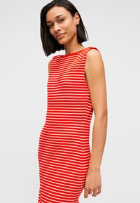 Νέα γυναίκα με κοντά σκούρα μαλλιά που φοράει αμάνικο φόρεμα με κόκκινες και λευκές ρίγες, στέκεται μπροστά σε μια απλή ανοιχτόχρωμη επιφάνεια.