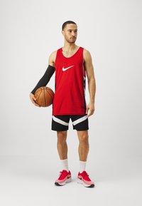 Red basketball tank top with white Nike logo, black shorts with white diagonal stripes, a black arm sleeve, and red athletic shoes.