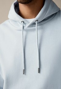 Helleblaue Kapuzenjacke mit Kordelzug. Weicher Stoff, mit Metallelementen an den Kordeln. Schlichtes, minimalistisches Design.